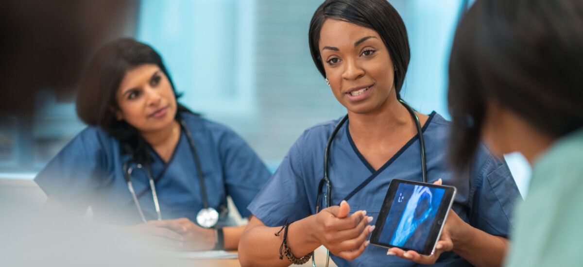 A Black female doctor leads a meeting with her medical team. She is showing the group a medical x-ray on a digital tablet. The multi-ethnic group of medical professionals is seated around a table in a conference room. They are discussing a treatment plan for their patient.