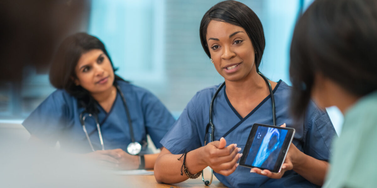 A Black female doctor leads a meeting with her medical team. She is showing the group a medical x-ray on a digital tablet. The multi-ethnic group of medical professionals is seated around a table in a conference room. They are discussing a treatment plan for their patient.