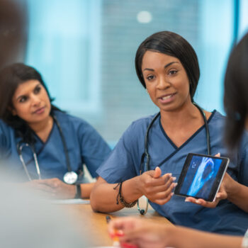 A Black female doctor leads a meeting with her medical team. She is showing the group a medical x-ray on a digital tablet. The multi-ethnic group of medical professionals is seated around a table in a conference room. They are discussing a treatment plan for their patient.