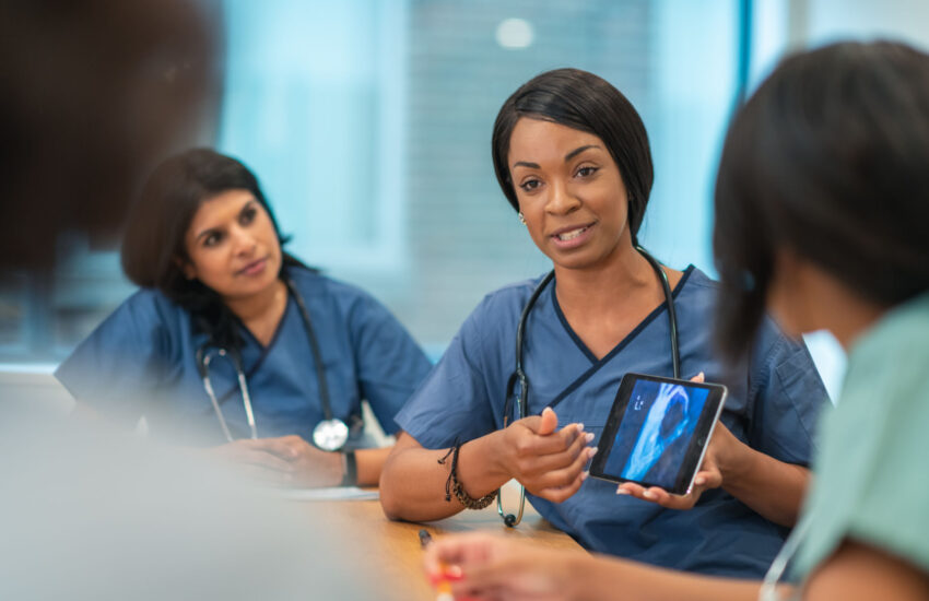 A Black female doctor leads a meeting with her medical team. She is showing the group a medical x-ray on a digital tablet. The multi-ethnic group of medical professionals is seated around a table in a conference room. They are discussing a treatment plan for their patient.